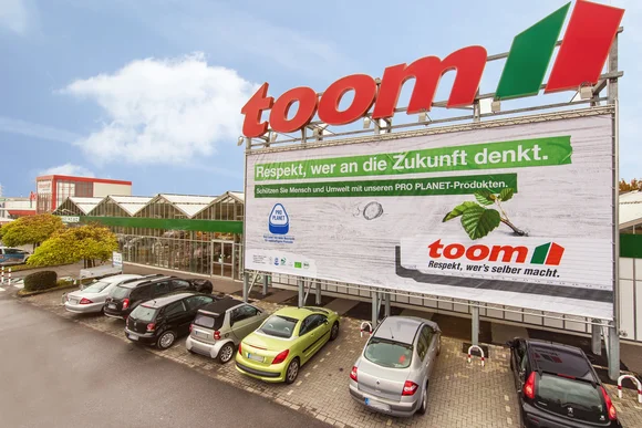 Fassade des toom Baumarktes mit Parkplatz und Plakat.Auf dem Plakat steht, Respekt wer an die Zukunft denkt. Schützen Sie Mensch und Umwelt mit unseren ProPlanet Produkten. Und das toom Logo. Respekt wer es selber macht.